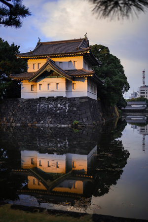 A historic Japanese castle gate sits on a stone moat, bathed in warm sunset light. Reflections ripple in the calm water, enhancing the tranquil scene of traditional architecture and surrounding trees.の写真素材