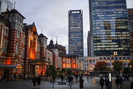 A bustling city square pairs a historic redbrick station with towering glass skyscrapers. Pedestrians fill the plaza as warm lights glow, blending heritage charm with urban energy.の写真素材