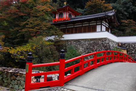 Serene autumn scene featuring a vibrant red arched bridge leading to traditional Japanese temple...