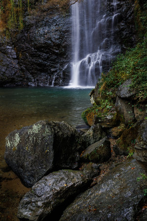 A serene autumn forest features a tall waterfall cascading over dark rocks, surrounded by colorful...