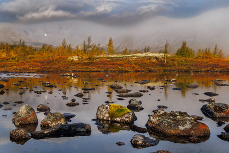 A tranquil autumn lakeside landscape with reflective water, scattered mossy rocks, and a distant...