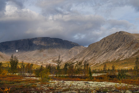 Broad valley framed by rugged mountains beneath heavy clouds. Autumn colors light the scattered...