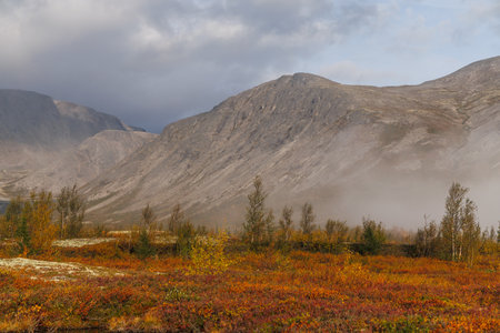Broad valley framed by rugged mountains beneath heavy clouds. Autumn colors light the scattered...