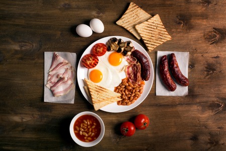 English breakfast prepared with two fried eggs, beans in tomato sauce, grilled tomatoes, mushrooms, bacon and toasts. Meal on a rustic wooden tableの写真素材