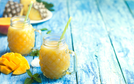 Fresh pineapple and mango smoothie in two glasses with fruits on a torquise wooden rustic backgroundの写真素材