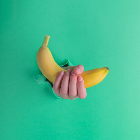 A woman holds a banana in her hand, inserted through a hole in torn green paperの写真素材