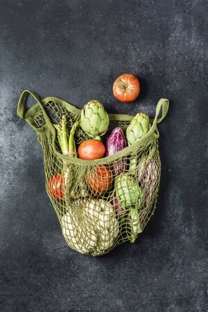 Fresh vegetables in a green string bag on a black background. Cauliflower, tomatoes, artichokes, asparagus and zucchini. No plastic, only natural materials. Top view. Copy space. Vertical orientation.の写真素材