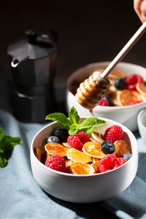 Tiny cereal small pancakes with blueberries, raspberries, mint and honey for a breakfast on a dark backgroundの写真素材