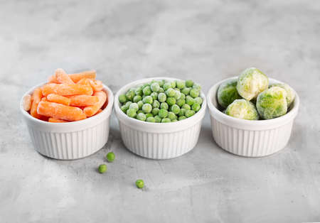 Frozen vegetables such as green peas, brussels sprouts and baby carrot in the white bowls on the concrete gray backgroundの写真素材