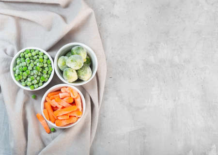 Frozen vegetables such as green peas, brussels sprouts and baby carrot in the white bowls on the concrete gray background with copy space, top viewの写真素材