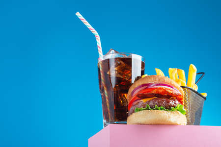 Fresh beef hamburger, cola and fried french fries placed on the pink stand and blue background. Copy space for text, trendy hero view. Horizontal orientationの写真素材