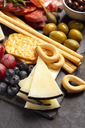 Charcuterie board with hard cheese, jamon, pork sausage with pepper, fuet, berries on a dark...