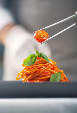 A chef in a white uniform and gloves serves cherry tomatoes in pasta in tomato sauce with basil leaves with tweezers . Haute cuisine. Plating foodの写真素材