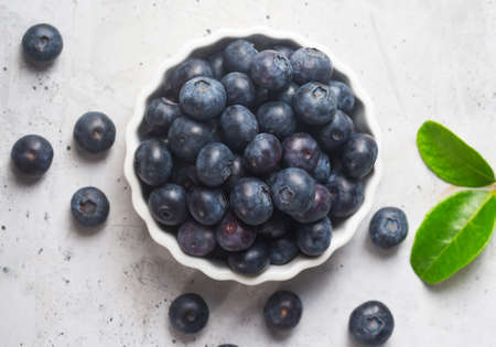 Organic ripe blueberries in a white bowl and scattered on a gray concrete background with a leaf top viewの写真素材