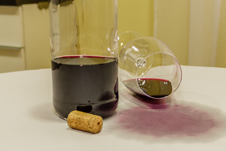 Red wine spilled from glass over white tablecloth with bottle and corkの写真素材