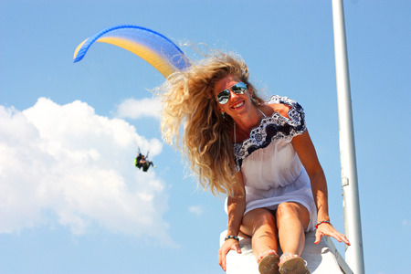 cheerful blond woman sitting in a white dress. Behind flies by a paragliderの写真素材
