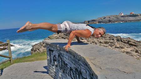 man in white shorts and white t-shirt does gymnastic exercise on a stone wall against a blue sea in...