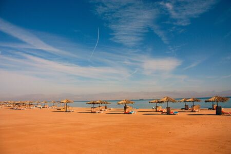 straw umbrellas from the sun on a beach on the shore of the red sea against a blue sky with white...
