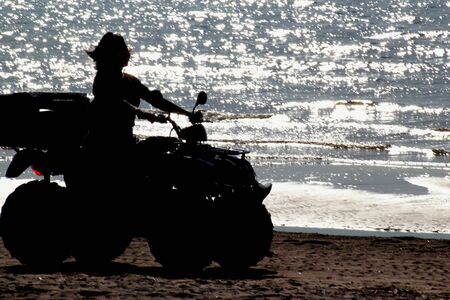 silhouette of a girl with long hair on the quad bike riding on the sand by the sea. Against the...