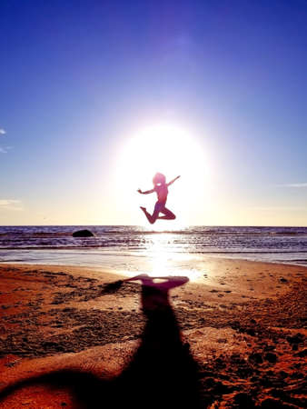 silhouette of a girl on the background of the sun disk in a jump on the sandy seashore in an interesting poseの写真素材