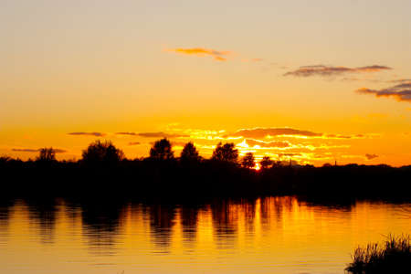 silhouette of trees on the river bank against the background of a yellow sunset. The sun in the clouds is reflected in the water of the riverの写真素材