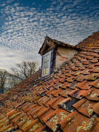 This captivating image showcases a neglected roof window of an old house against the blue sky. The wooden window adds character to the weathered roof with red tiles, creating a nostalgic scene.の写真素材
