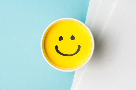 Yellow emoticon smiley face or happy emotion, on paper cup and papers over blue background.の写真素材