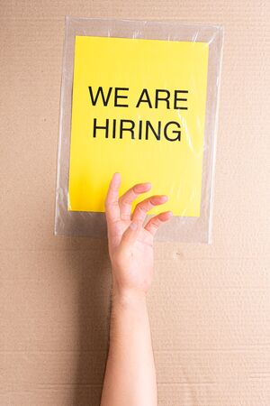Hand raised trying to reach a classified ad. We are hiring printed on yellow paper on transparent plastic bag with cardboard background. Job aspirations, opportunity, search conceptの写真素材