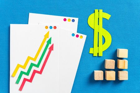 Inflation and financial concepts as profitability, taxes, stock market. Three paper arrows growing up and paper green dollar symbol with graph bar made with wooden blocks, on blue textured background.の写真素材