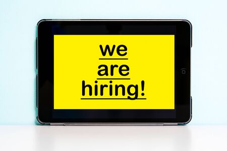 Yellow note with We are hiring phrase on screen tablet pc and blue background. Job search, ...