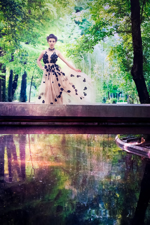 Beautiful female model with long tulle flower dress and big hair standing on a bridge over colorful and bright water in a park. Fine art conceptの写真素材
