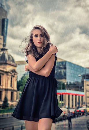 Beautiful blonde girl with big blue eyes and black dress, feeling cold and covering herself, during rain, in the city, on the street, during daytimeの写真素材