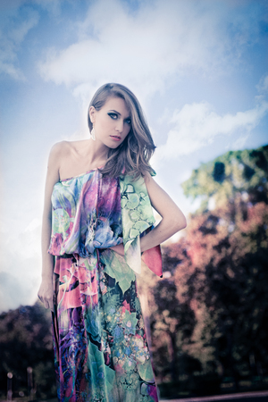 Beautiful blonde woman with big blue eyes, looking straight, wearing a silk colorful dress with bare shoulder, in the park, during daytimeの写真素材