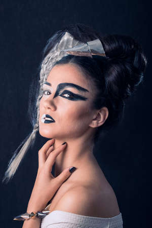 Beautiful Caucasian brunette close-up, with half face painted with white make-up and half wih graphic black make-up. Her hair is half white with a wing made of braided hair and half black with a silver horn. She is scratching her neck, on black background. Angel and Devil Conceptの写真素材