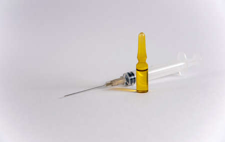 Dark yellow glass ampoule with liquid stands in front of a lying medical syringe on a white...