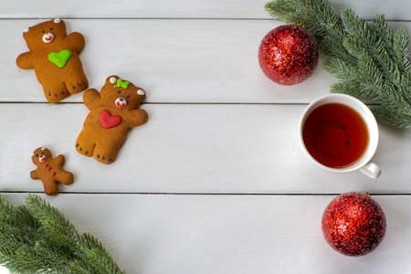 Layout on a white wooden background of three cookies in the form of bears, artificial fir branches, mugs of tea and red shiny Christmas balls top viewの写真素材