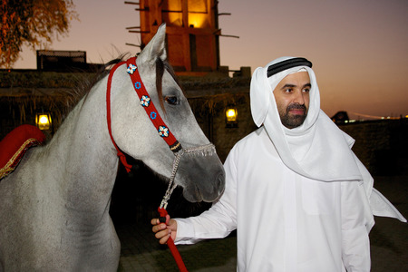 An Arab man leads his grey Arabian horse at sunset in Dubai. Traditional paraffin lamps and a windtower can be seen in the background.のeditorial素材