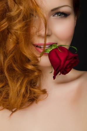 young woman with long red hair holds beautiful rose in her mouthの写真素材