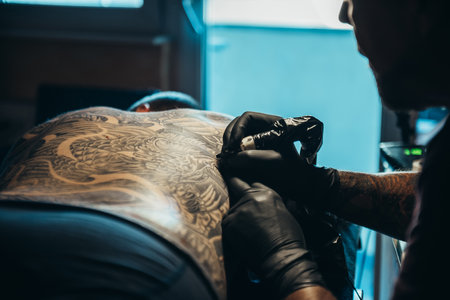 Hands of a tattoo artist wearing black gloves and holding a machine while creating a picture on a man backの写真素材