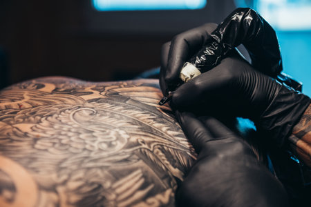 Hands of a tattoo artist wearing black protective gloves creating a picture on a man back while...