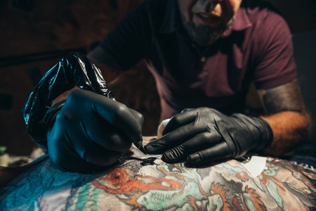 Tattoo artist wearing black gloves creating a picture on a mans back with a tattoo machineの写真素材