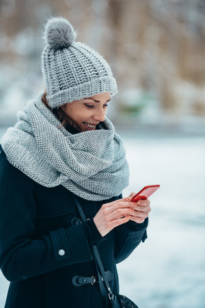 Beautiful young woman using smartphone and wearing scarf and a a hat on a cold winter day during snowの写真素材
