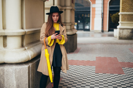 Young beautiful woman using a smartphone and holding a yellow umbrella while walking outside on a rainy dayの写真素材