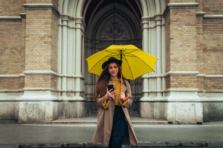 Young beautiful woman using a smartphone and holding a yellow umbrella while walking in a cit on a...