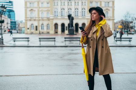 Young beautiful woman using smartphone and holding a yellow umbrella while out in the cityの写真素材