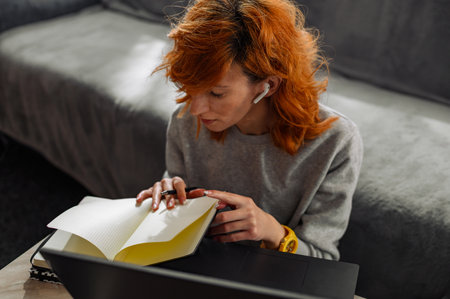 Young redhead woman using a laptop, airpods and a notebook while working from home