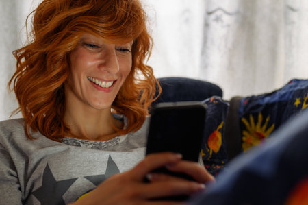 Young redhead woman using a smartphone and lying in bedsheets while being illuminated by the device...