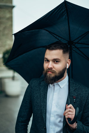 Young handsome man using a  while holding a black umbrella while walking in a city on a rainy dayの写真素材