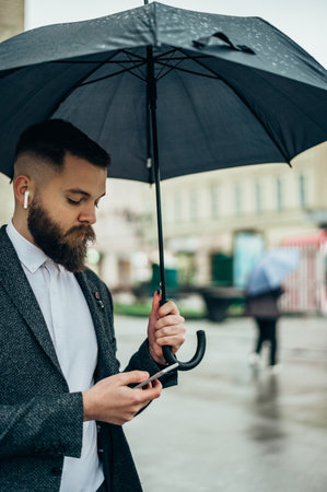 Young handsome man using a smartphone while holding a black umbrella while walking in a city on a rainy dayの写真素材
