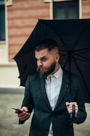 Young handsome businessman using a smartphone and holding a black umbrella while outside on a rainy dayの写真素材